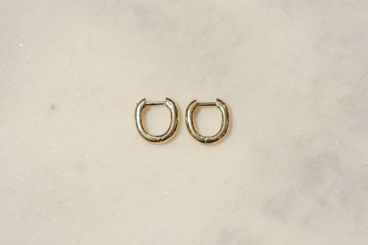 These earrings are gold-toned hoops with a unique, slightly irregular shape. They have a smooth, polished surface that reflects light, giving them a shiny appearance. The hoops are slightly flattened and are of medium thickness. They feature a simple hinge and latch closure mechanism. The overall design is minimalistic yet elegant, suitable for both everyday wear and more formal occasions.