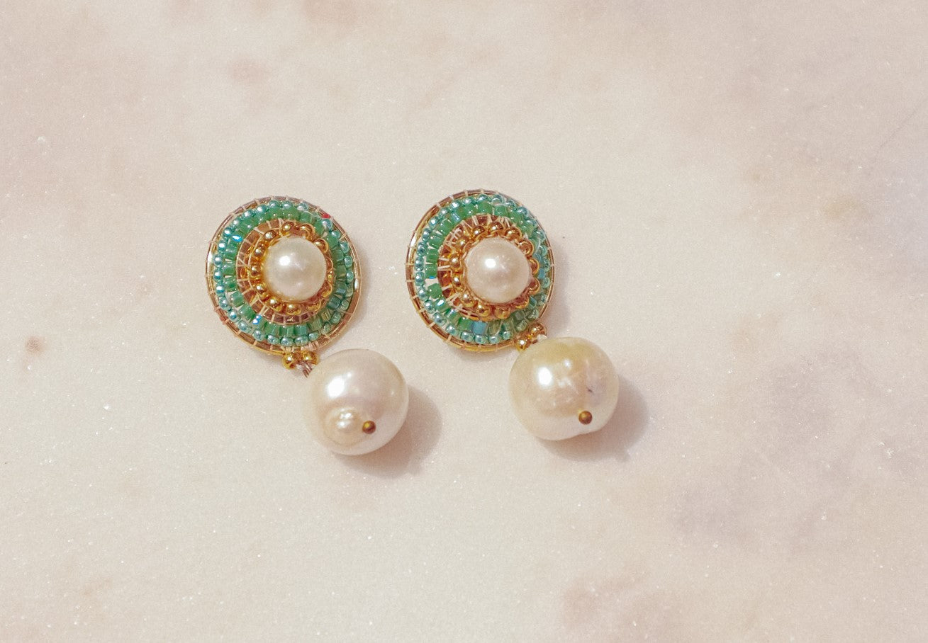 These earrings showcase a captivating interplay of classic and modern design. Luminous pearl drops exude timeless elegance, while shimmering green Miyuki beads, meticulously woven around the posts, add a touch of modern flair. Secured on gleaming 18k gold-filled posts, these earrings are perfect for everyday wear or a touch of elegance for a special occasion