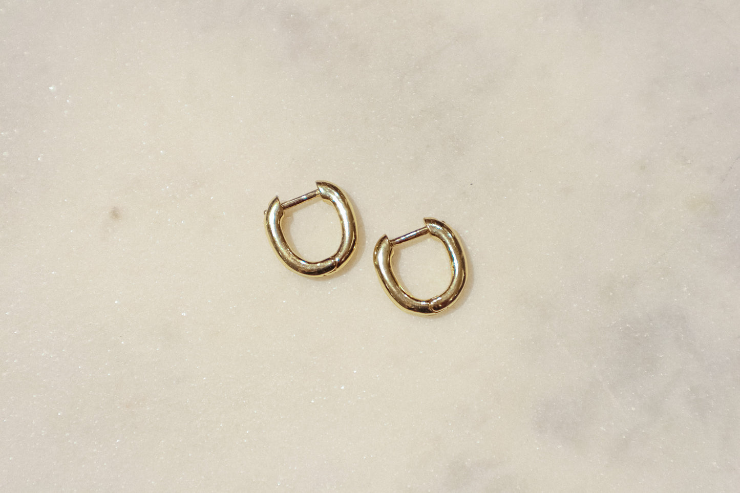 These earrings are gold-toned hoops with a unique, slightly irregular shape. They have a smooth, polished surface that reflects light, giving them a shiny appearance. The hoops are slightly flattened and are of medium thickness. They feature a simple hinge and latch closure mechanism. The overall design is minimalistic yet elegant, suitable for both everyday wear and more formal occasions.