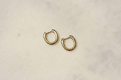 These earrings are gold-toned hoops with a unique, slightly irregular shape. They have a smooth, polished surface that reflects light, giving them a shiny appearance. The hoops are slightly flattened and are of medium thickness. They feature a simple hinge and latch closure mechanism. The overall design is minimalistic yet elegant, suitable for both everyday wear and more formal occasions.