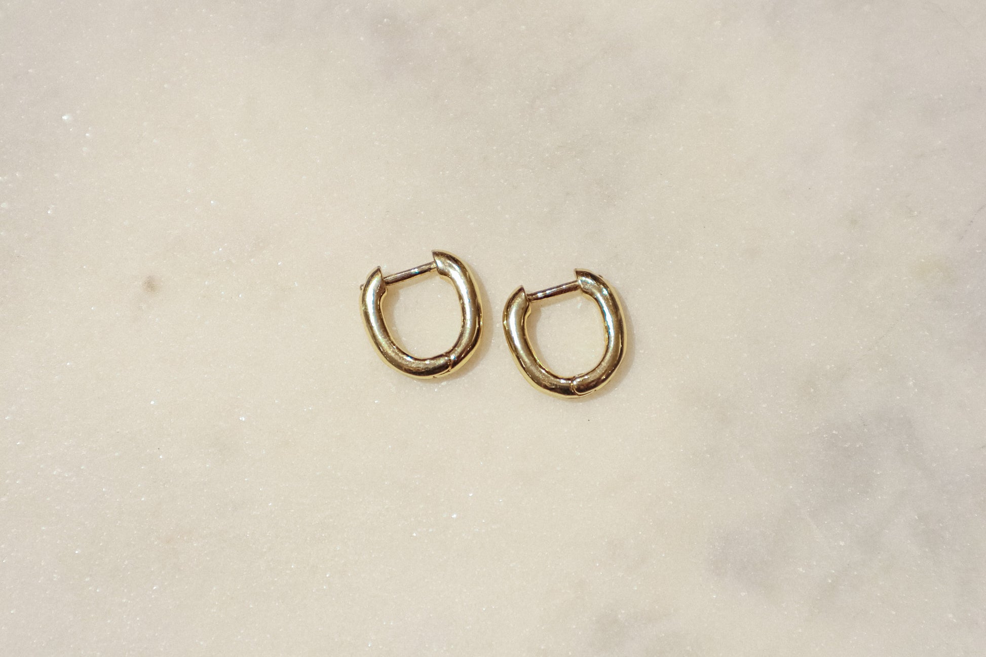 These earrings are gold-toned hoops with a unique, slightly irregular shape. They have a smooth, polished surface that reflects light, giving them a shiny appearance. The hoops are slightly flattened and are of medium thickness. They feature a simple hinge and latch closure mechanism. The overall design is minimalistic yet elegant, suitable for both everyday wear and more formal occasions.