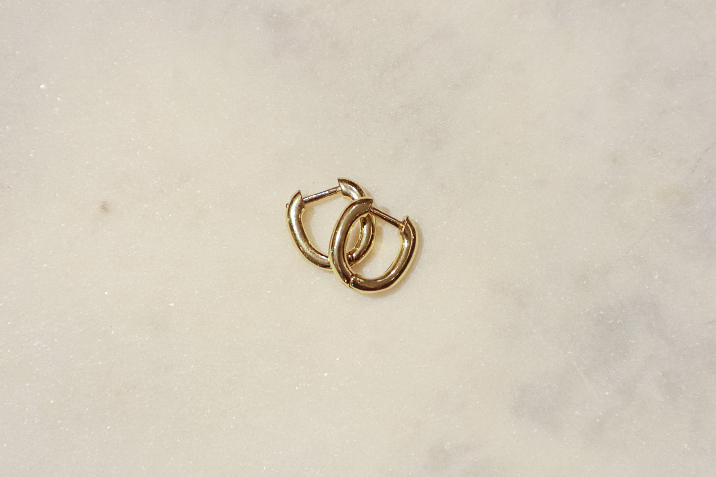 These earrings are gold-toned hoops with a unique, slightly irregular shape. They have a smooth, polished surface that reflects light, giving them a shiny appearance. The hoops are slightly flattened and are of medium thickness. They feature a simple hinge and latch closure mechanism. The overall design is minimalistic yet elegant, suitable for both everyday wear and more formal occasions.