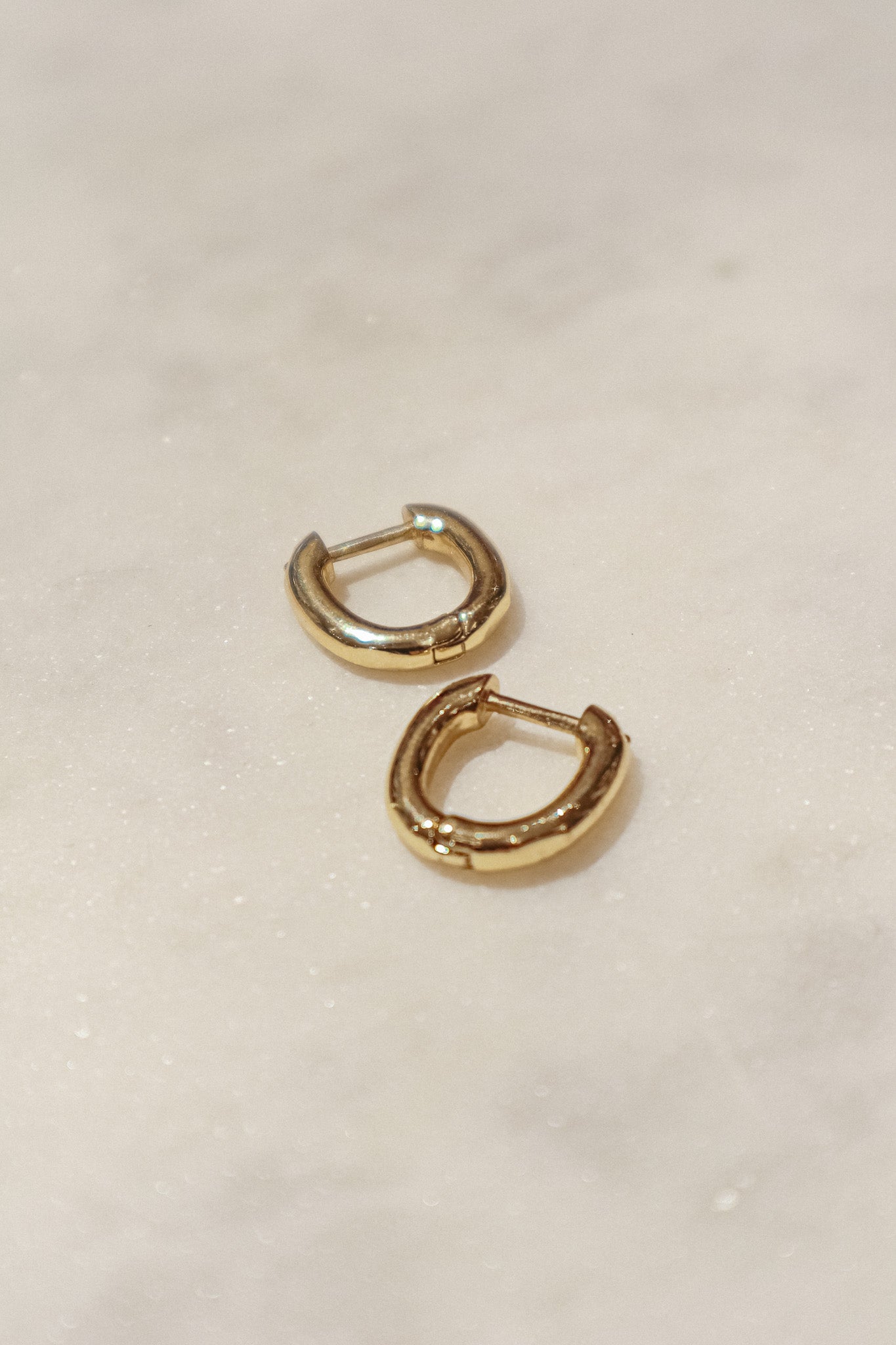 These earrings are gold-toned hoops with a unique, slightly irregular shape. They have a smooth, polished surface that reflects light, giving them a shiny appearance. The hoops are slightly flattened and are of medium thickness. They feature a simple hinge and latch closure mechanism. The overall design is minimalistic yet elegant, suitable for both everyday wear and more formal occasions.