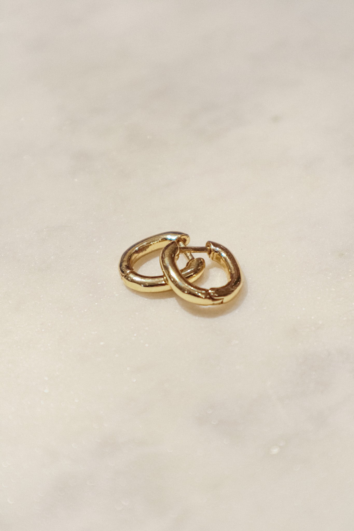 These earrings are gold-toned hoops with a unique, slightly irregular shape. They have a smooth, polished surface that reflects light, giving them a shiny appearance. The hoops are slightly flattened and are of medium thickness. They feature a simple hinge and latch closure mechanism. The overall design is minimalistic yet elegant, suitable for both everyday wear and more formal occasions.